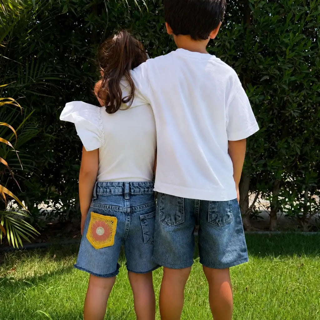 Two children standing outdoors on grass with bushes behind them, viewed from behind. The child on the left wears a white puff-sleeve top and blue denim shorts with a yellow flower patch pocket, and the child on the right wears a white T-shirt and matching blue denim shorts.