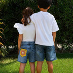 Two children standing outdoors on grass with bushes behind them, viewed from behind. The child on the left wears a white puff-sleeve top and blue denim shorts with a yellow flower patch pocket, and the child on the right wears a white T-shirt and matching blue denim shorts.