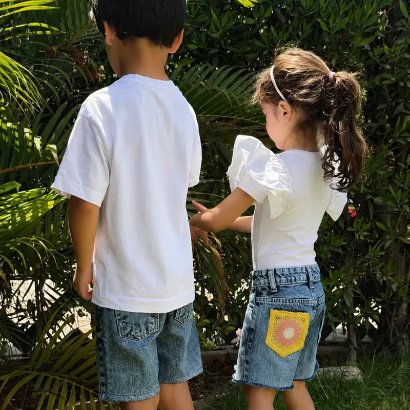 Two children standing outdoors in front of green foliage, wearing white short-sleeve tops and denim shorts. The child on the right has a yellow patchwork detail on the back of their denim shorts.