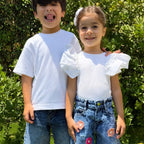 Two children standing outdoors in front of green foliage. The boy wears a plain white short-sleeve T-shirt and blue jeans, while the girl wears a white short-sleeve top with puffed ruffled sleeves and light blue jeans with flower patches.