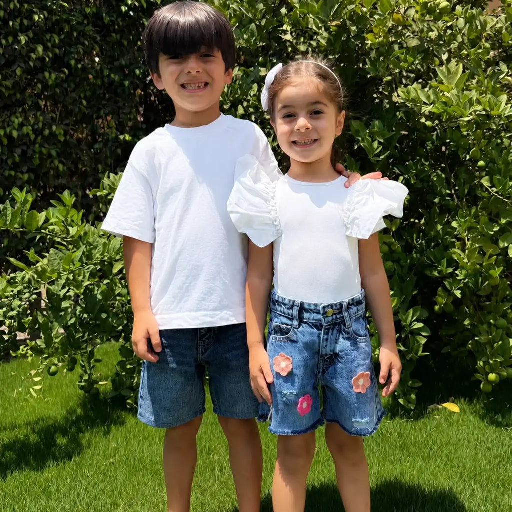 Two children standing outdoors on a grassy lawn in front of a green hedge. The boy wears a plain white short-sleeve T-shirt and blue denim shorts, while the girl wears a white short-sleeve top with puffed ruffle sleeves and distressed blue denim shorts with floral patches.