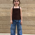 Child wearing a brown top and blue jeans standing against a wooden wall.