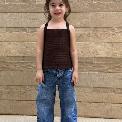 Child wearing a brown top and blue jeans standing against a wooden wall.