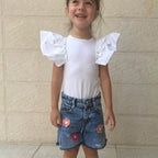A young girl smiling and wearing a white short-sleeve top with dramatic ruffled flutter sleeves, paired with blue denim shorts featuring pink flower appliqués, standing against a beige textured wall.