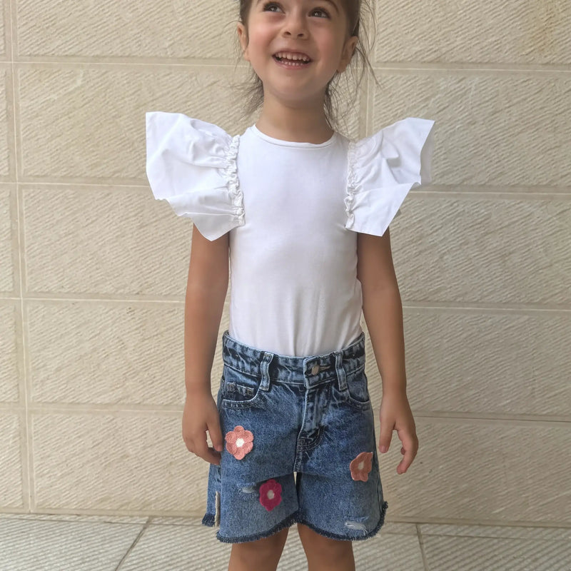A young girl smiling and wearing a white short-sleeve top with dramatic ruffled flutter sleeves, paired with blue denim shorts featuring pink flower appliqués, standing against a beige textured wall.