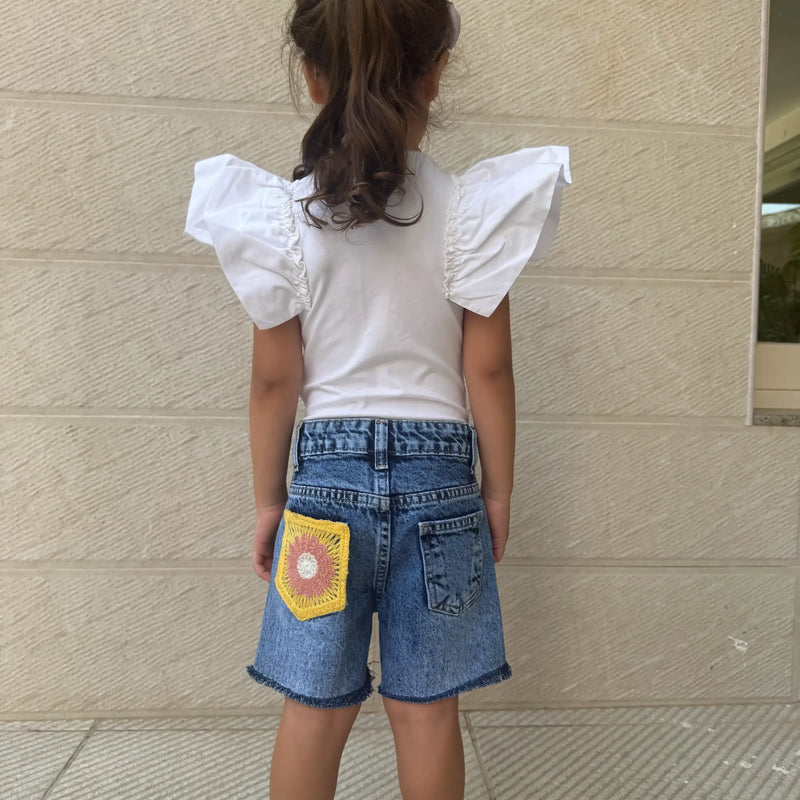A child viewed from behind wearing a white top with puffed flutter sleeves and blue denim shorts featuring a yellow crochet patch on the back pocket, standing against a beige textured wall.