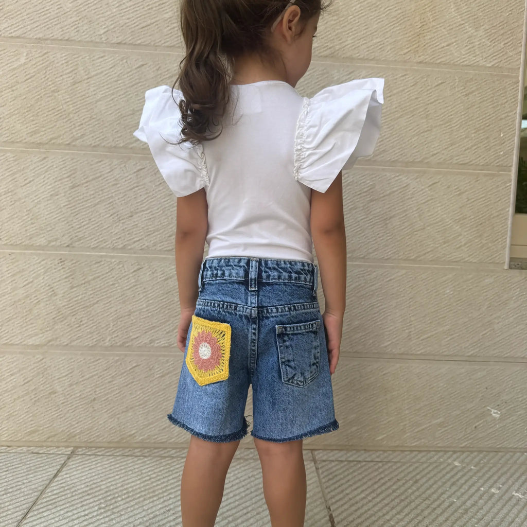 A young girl viewed from behind wearing a white short-sleeve top with dramatic ruffled shoulders and blue denim shorts featuring a yellow crocheted patch on the back pocket, standing against a beige textured wall.