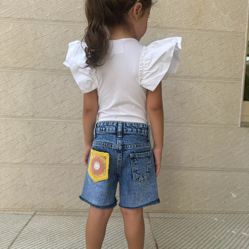 A young girl viewed from behind wearing a white short-sleeve top with dramatic ruffled shoulders and blue denim shorts featuring a yellow crocheted patch on the back pocket, standing against a beige textured wall.