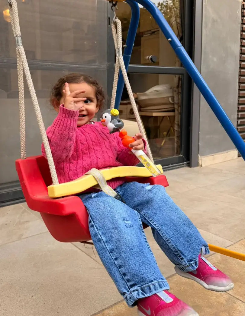 Child in a red swing set with a toy, sitting on a sidewalk.